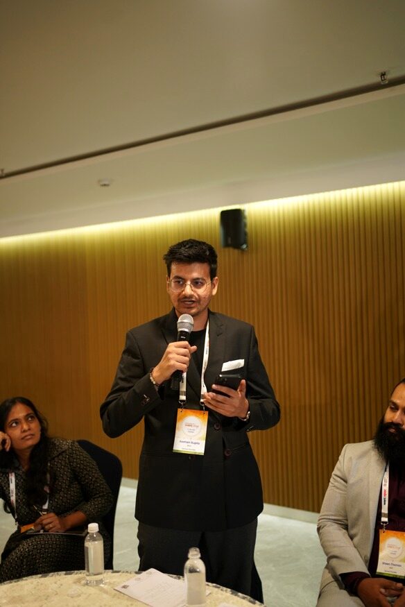 Eashan Gupta standing at the roundtable with a microphone, reporting the AI table's conclusions back to the room